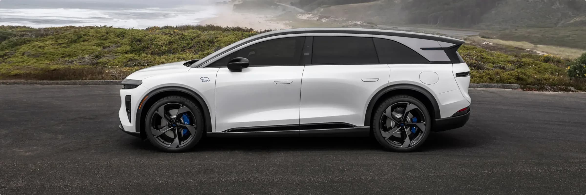 Lucid Gravity GT-S màu trắng với nóc đen và kẹp phanh xanh đỗ trên cung đường ven biển, phía sau là biển mù sương và những ngọn đồi xanh.
